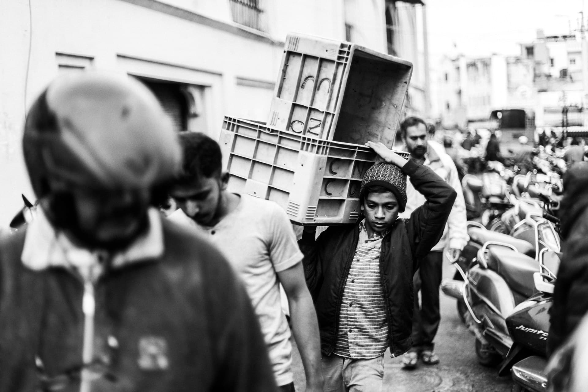 A young man carries a stack of plastic crates on his shoulder down a busy street lined with parked scooters. Black and white.