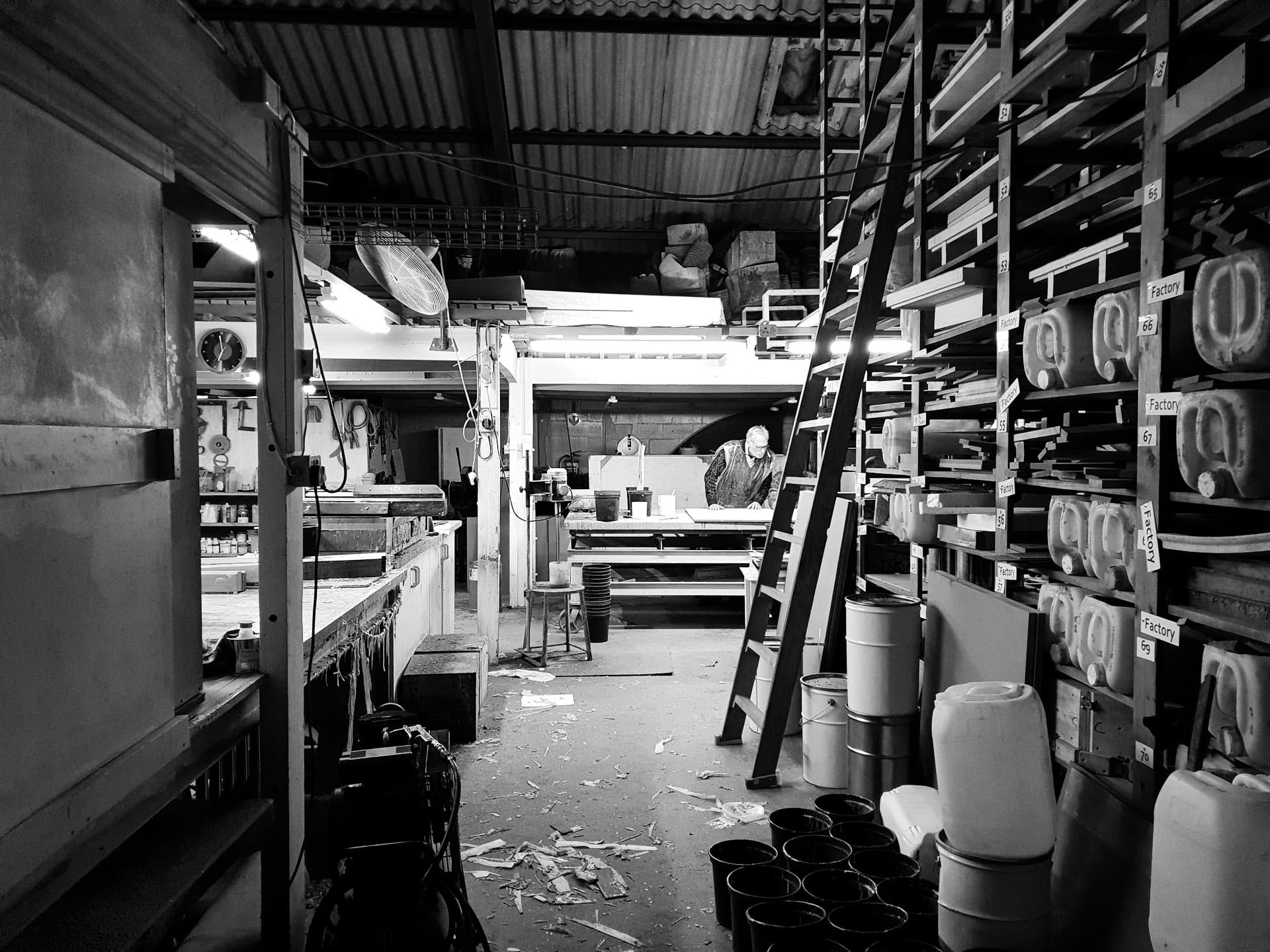 A long workshop aisle lined with shelves of containers and supplies, a man working at a bench at the far end. Black and white.