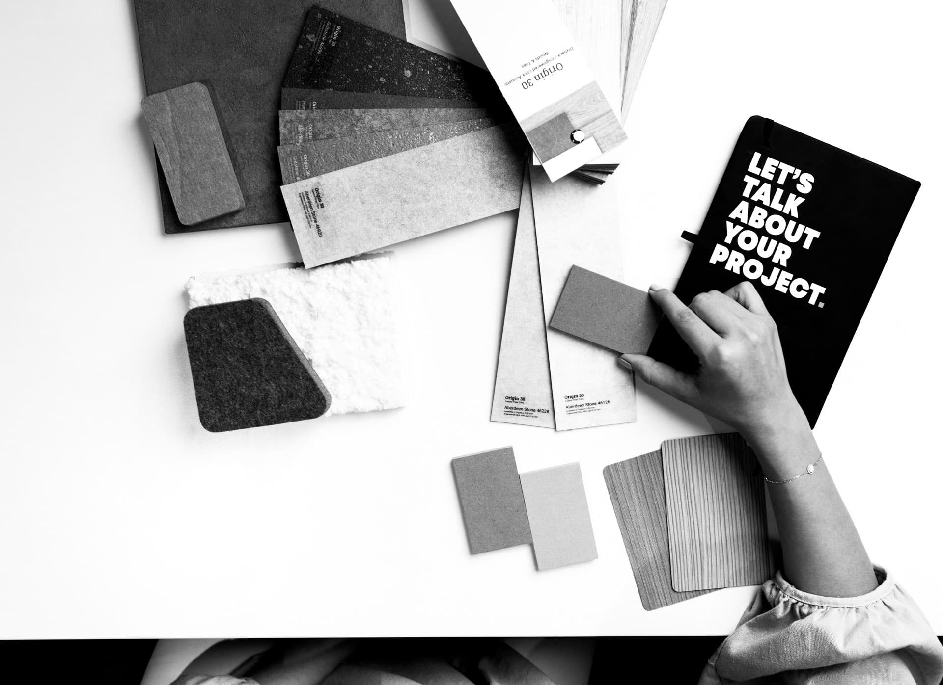 A designer’s hand arranges wood, stone, felt and laminate finish samples on a white table beside a notebook. Black and white.